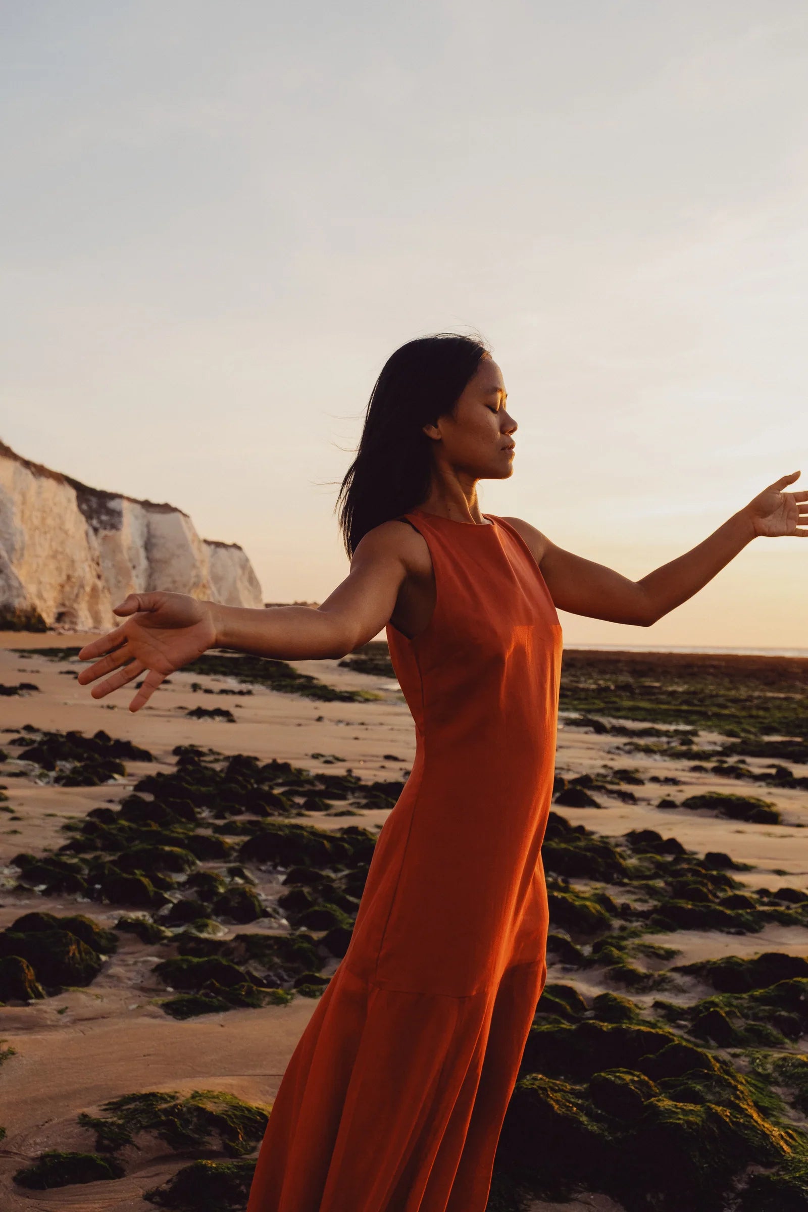 A person in a red dress with arms outstretched on a beach at golden hour.