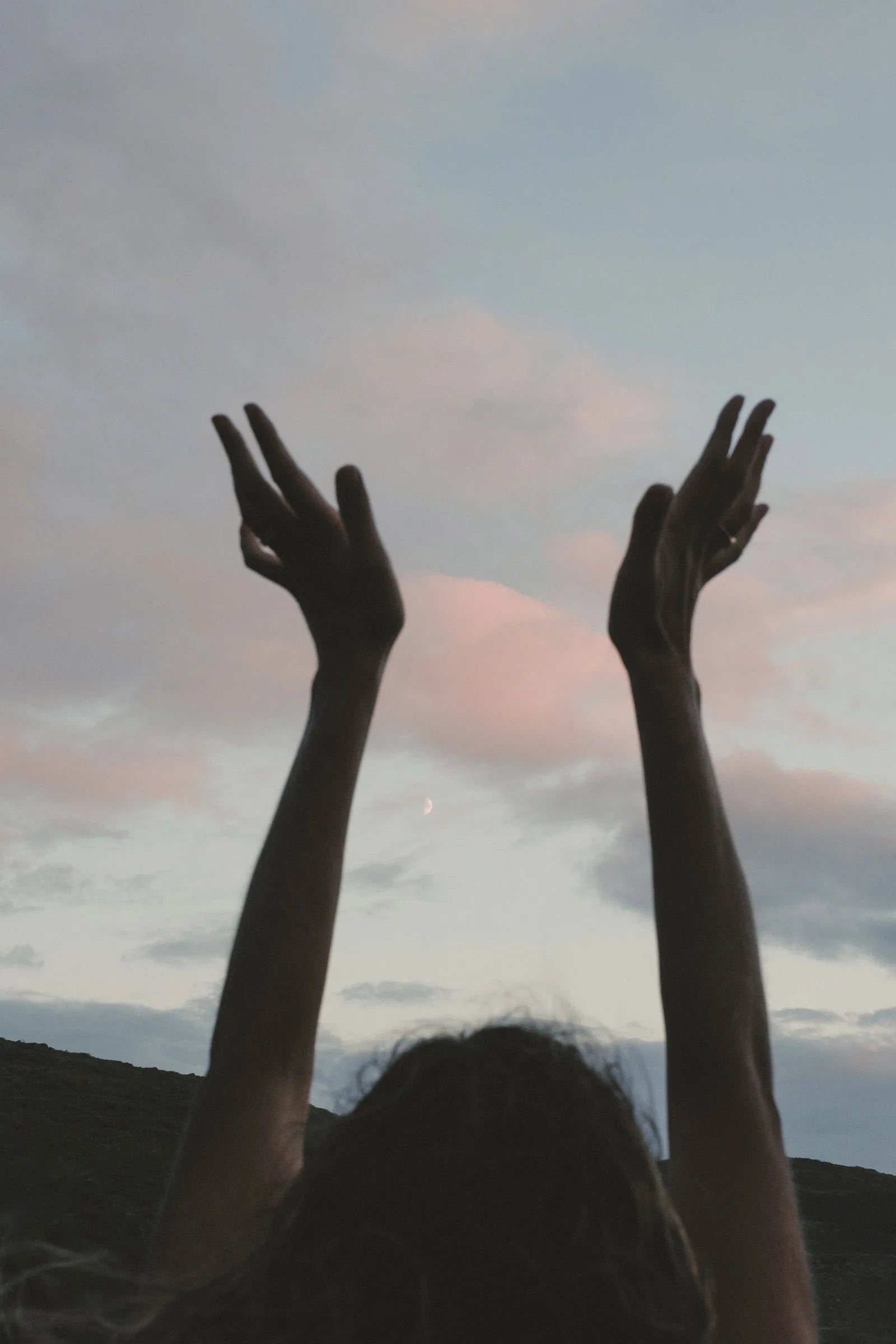 Hands reaching towards the moon in a soft evening sky.